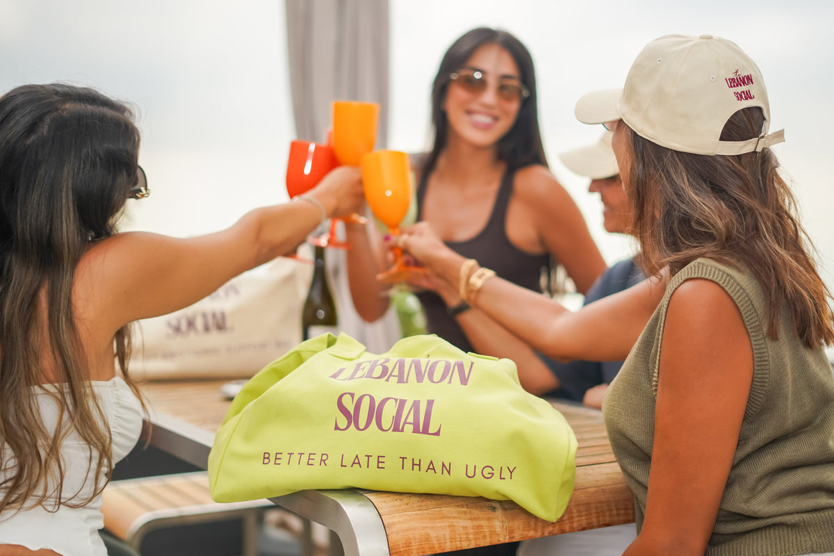 Three women sitting and clinking wine glasses at a table, with a bright lime green tote bag in the foreground that reads 'LEBANON SOCIAL BETTER LATE THAN UGLY'.