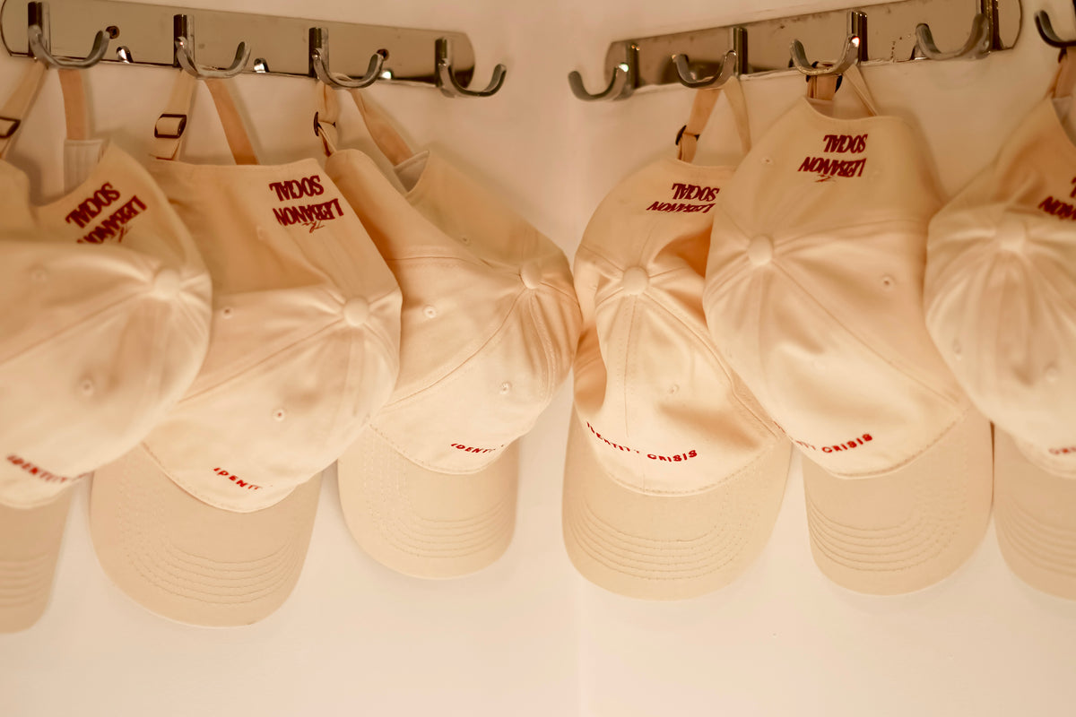 A row of cream-colored baseball caps hanging from metal hooks on a wall. Each cap features red embroidered text reading 'TOKYO MONARCH' on the front panels.